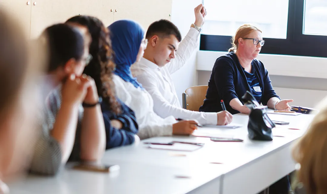 Mehrere Schüler:innen sitzen an einem Tisch im Klassenraum, hören zu und arbeiten, eine Person meldet sich.