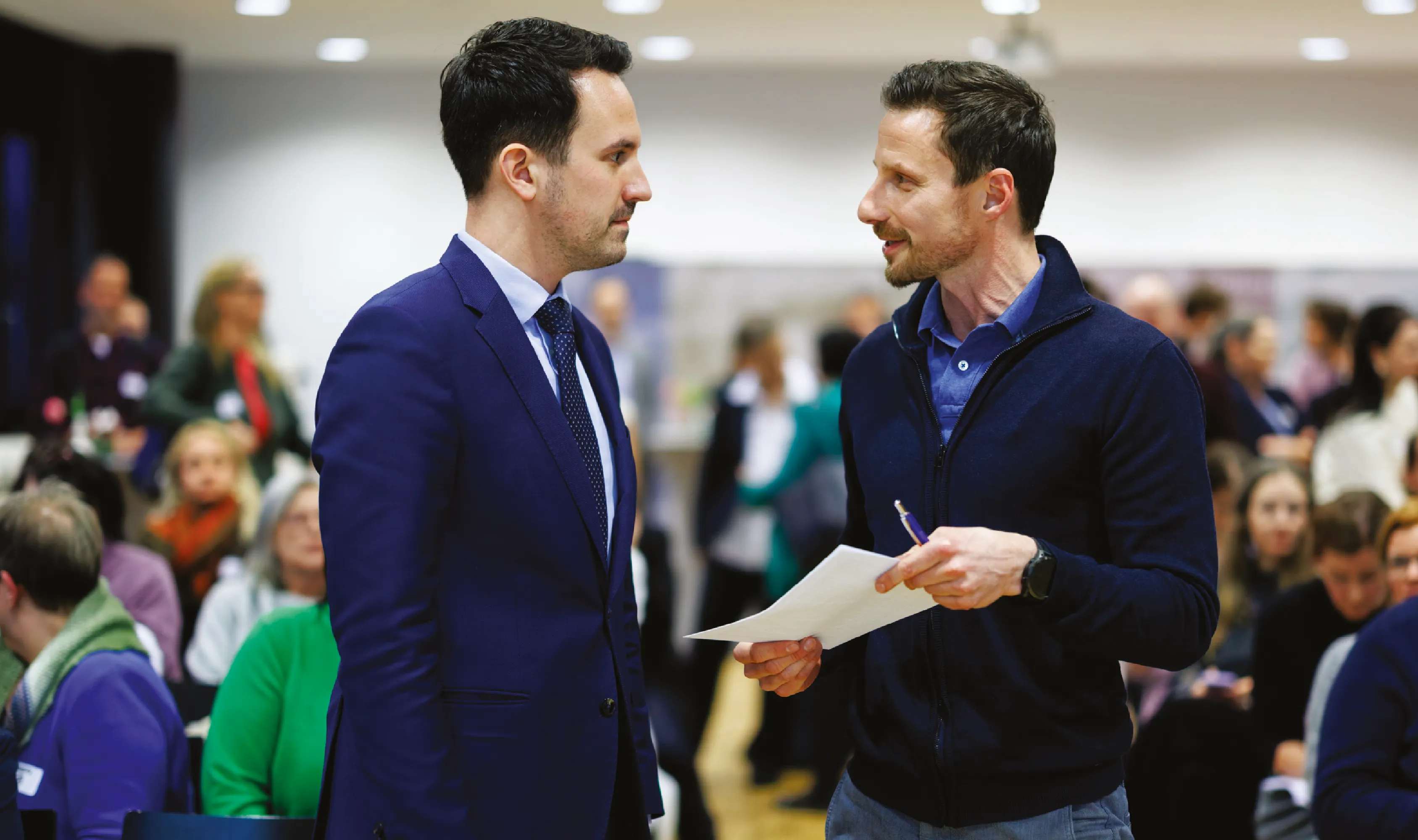 Christoph Wiederkehr im Gespräch mit einem Mann, der ein Blatt Papier hält, im Hintergrund eine größere Veranstaltung mit Publikum.