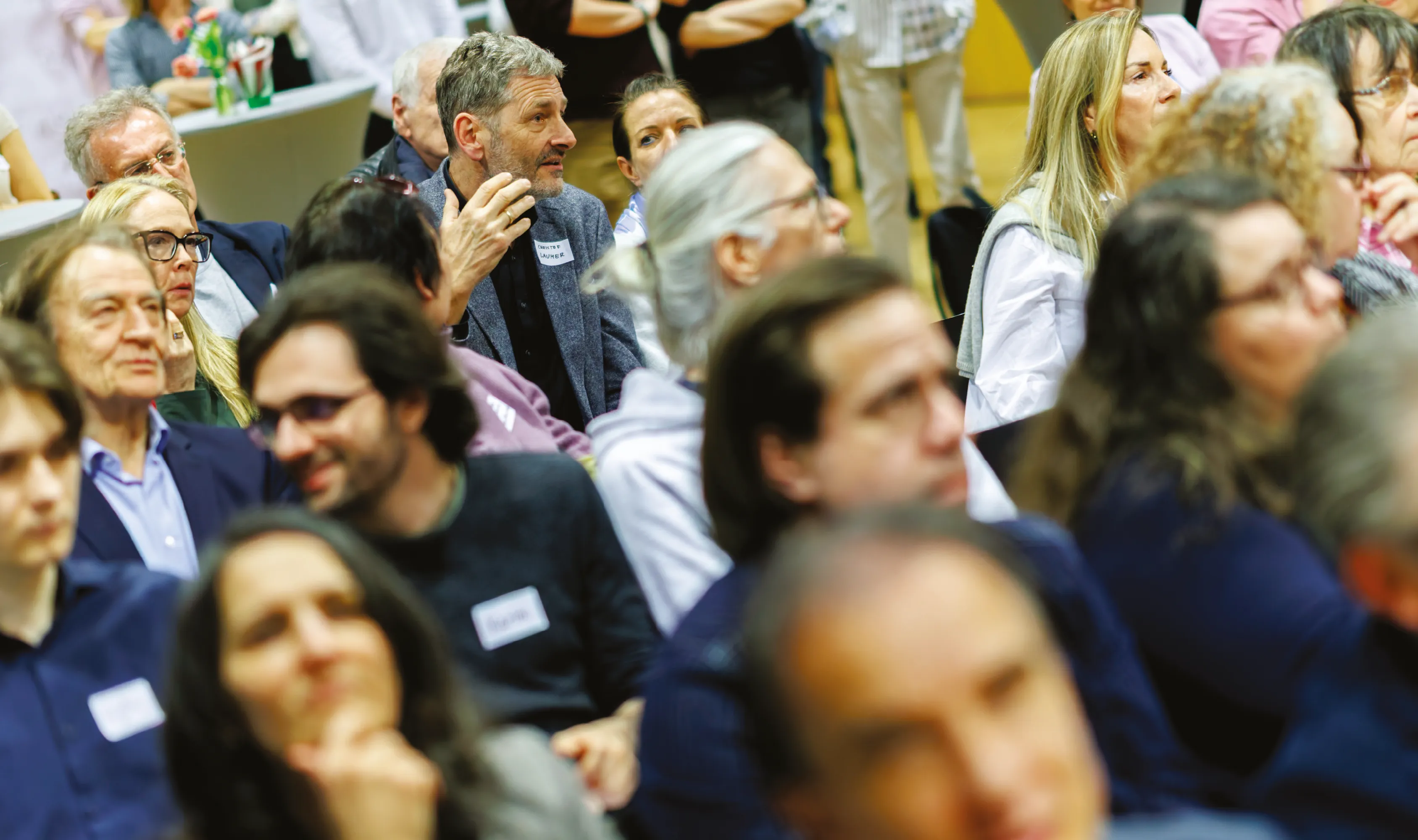 Ein Publikum aus Erwachsenen sitzt dicht beisammen und hört aufmerksam zu, einige melden sich oder diskutieren.