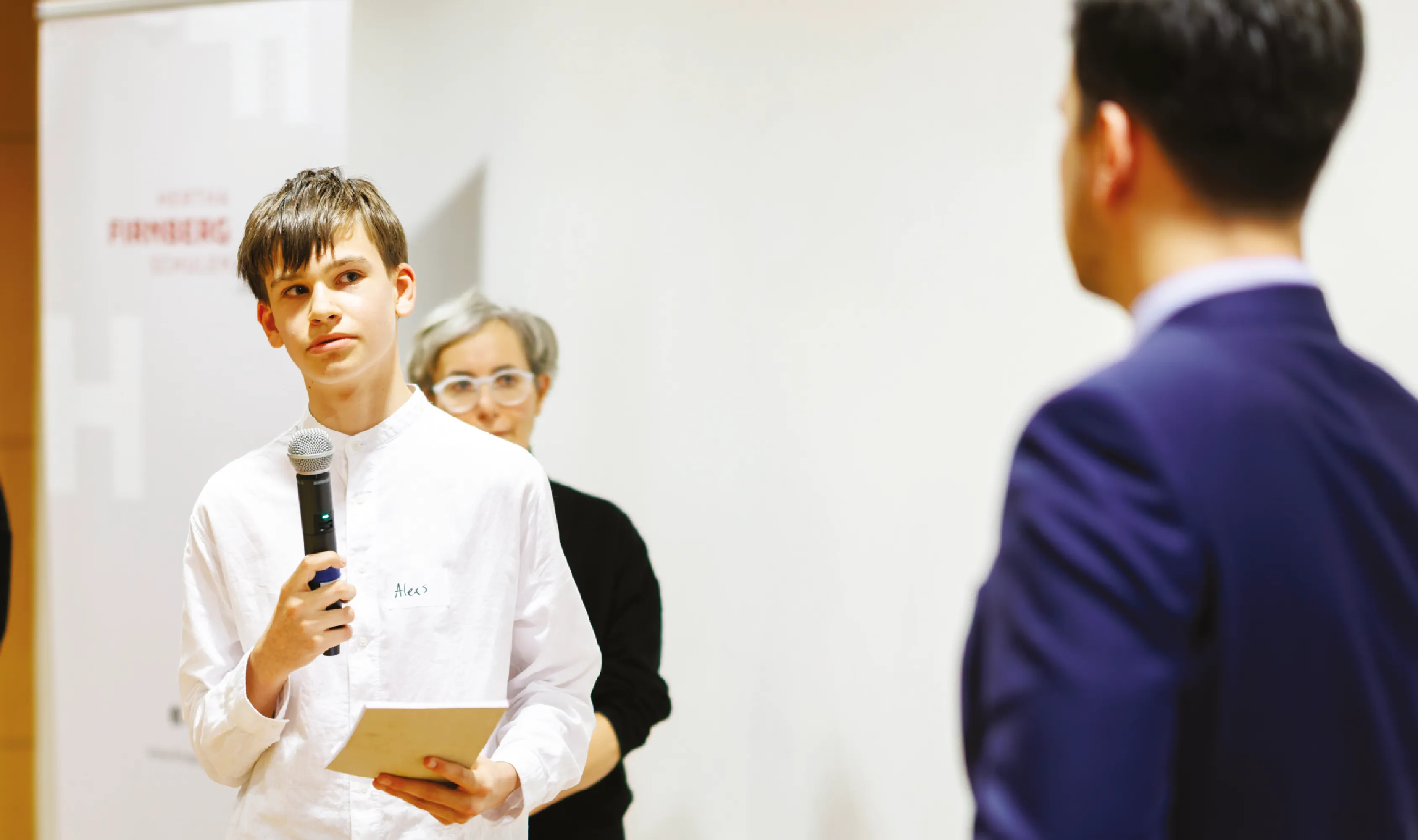 Ein Jugendlicher hält ein Mikrofon und stellt eine Frage oder macht einen Beitrag während einer Veranstaltung, während ein Mann im Anzug zuhört.
